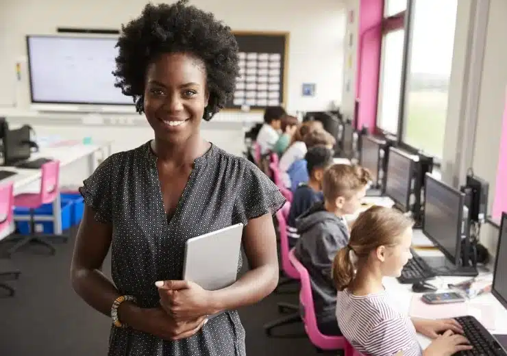 Female teacher in the middle of a computer class smiling for a photo while kids are using computers.